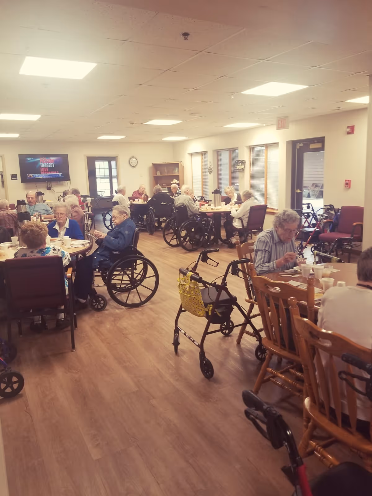 Elderly residents eating and socializing around tables in a bright assisted-living dining room with wheelchairs and walkers present.