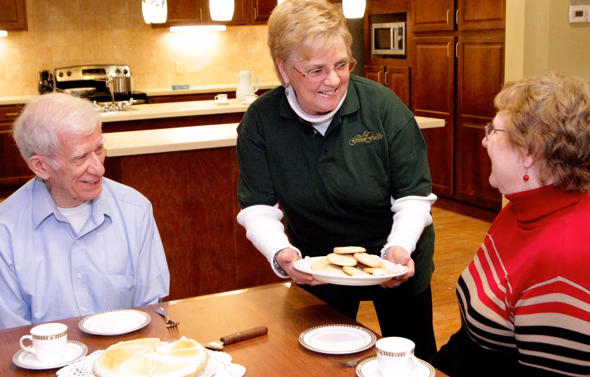 A staff member serves a plate of cookies to two smiling elderly residents seated at a dining table in a communal kitchen.