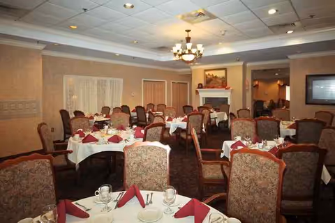 A dining room with multiple round tables covered with white tablecloths and set with plates, glasses, silverware, and folded red napkins. The room features upholstered chairs, a chandelier hanging from the ceiling, a fireplace with a painting above it, and warm beige walls.