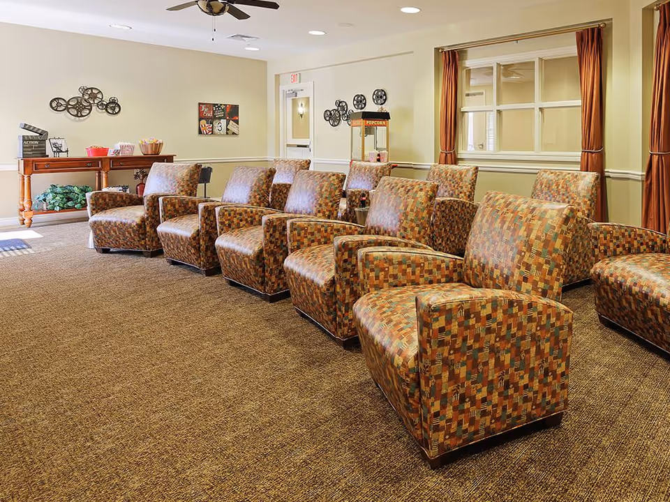 A cozy common area with multiple patterned armchairs arranged in rows facing a popcorn machine. The room has beige walls, a brown carpet, ceiling fans, and decorative wall art. A wooden table with popcorn containers and a clapperboard is visible in the background.