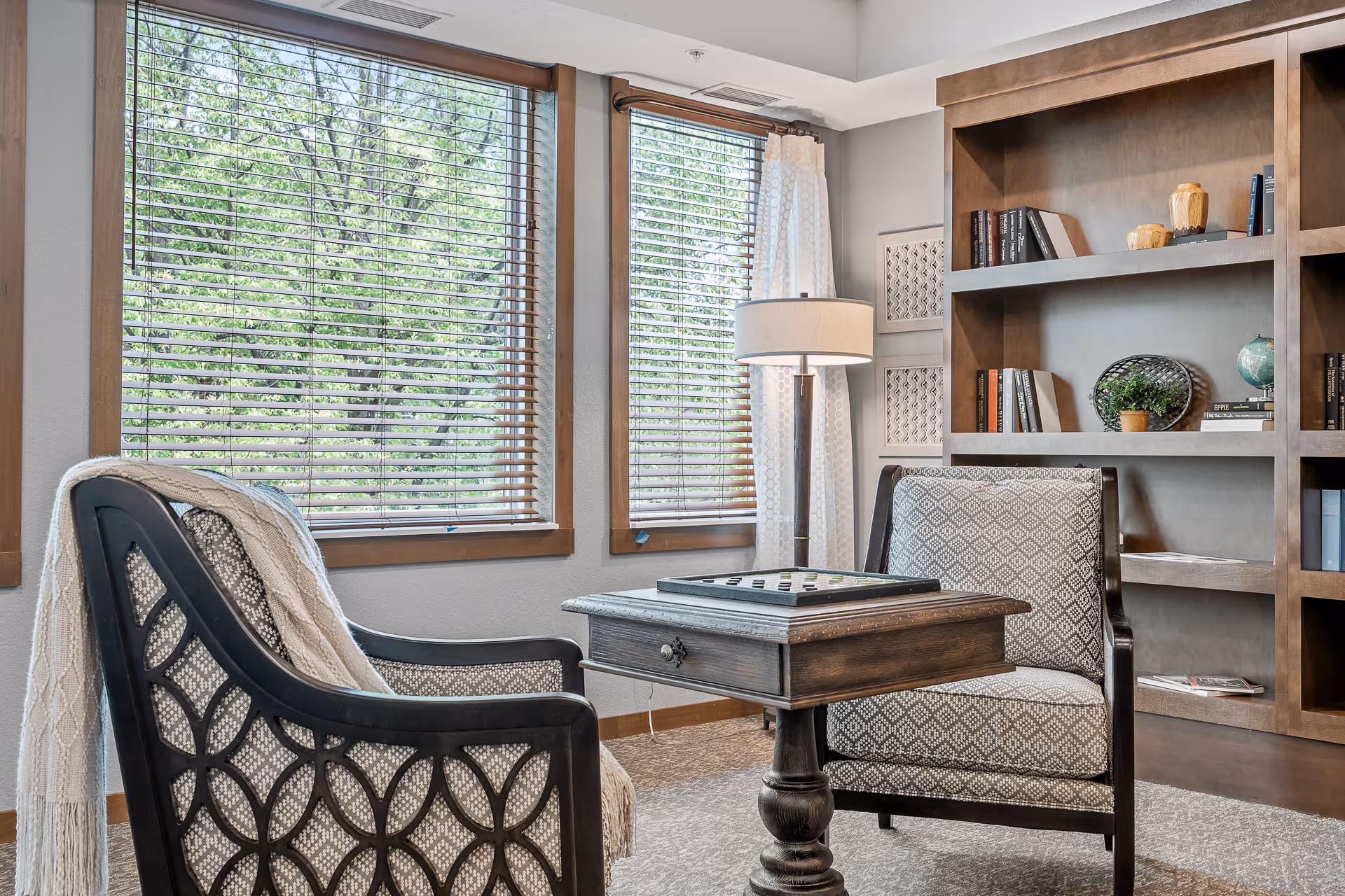 A cozy sitting area with two patterned armchairs facing a small wooden table with a game board on top. Behind one chair is a large window with wooden blinds and white curtains, letting in natural light. A tall floor lamp stands between the window and a wooden bookshelf filled with books and decorative items.
