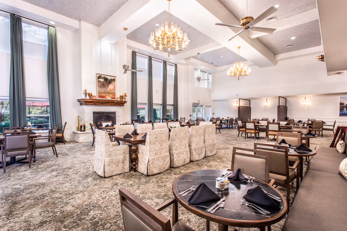 Spacious dining room with multiple tables and chairs arranged neatly. The room features high ceilings with chandeliers and ceiling fans, large windows with long curtains, and a fireplace with a painting above it. Tables are set with black napkins and silverware, creating an elegant atmosphere.