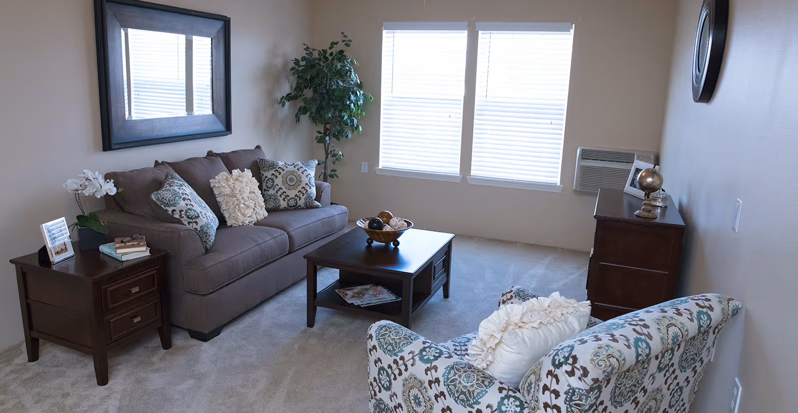 A cozy living room with a brown sofa adorned with decorative pillows, a patterned armchair with a white pillow, a dark wooden coffee table with a decorative bowl, a side table with books and a framed photo, a large mirror on the wall, a potted plant, two windows with blinds, and a wooden dresser with a globe and framed photo.