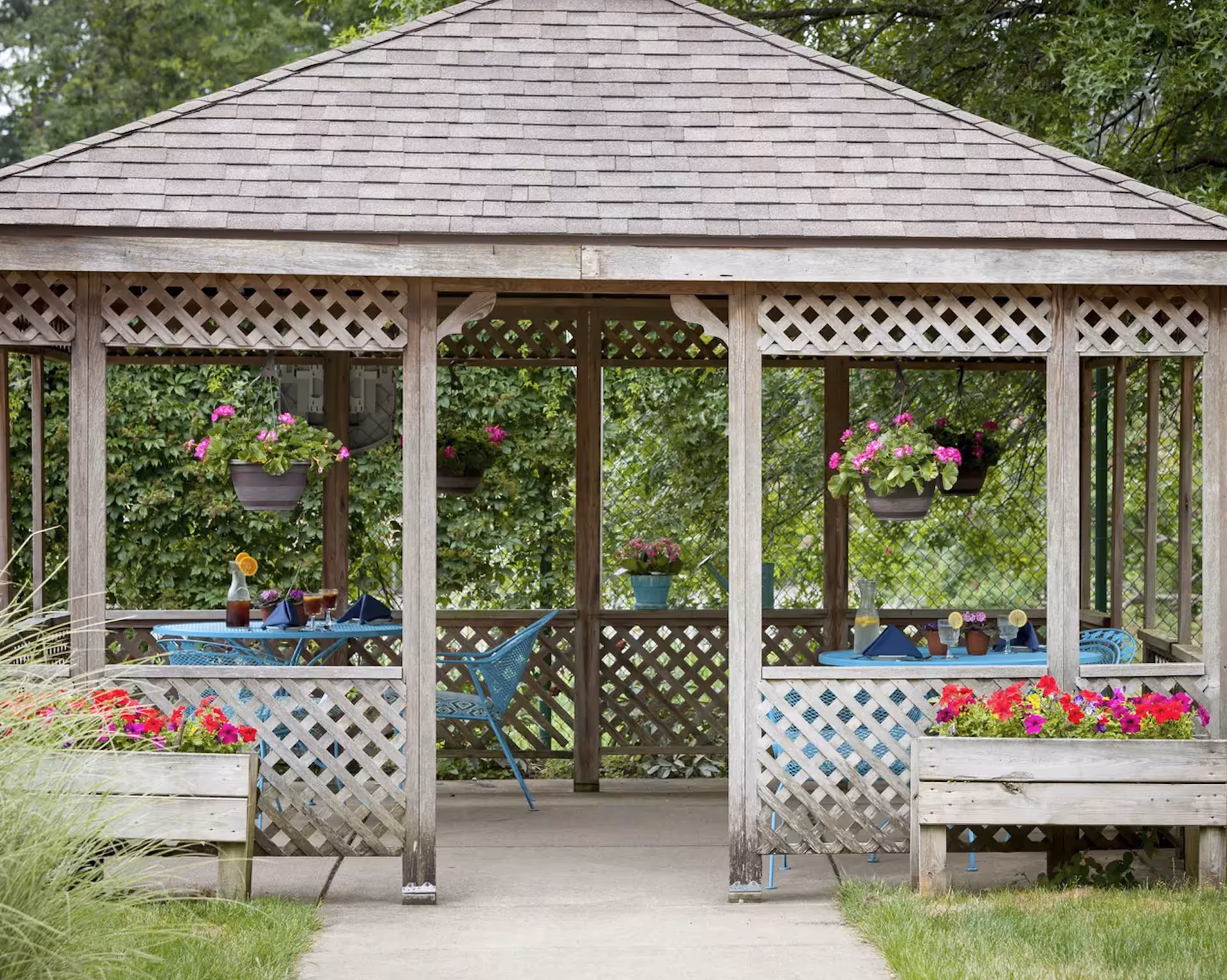 A wooden gazebo with lattice sides, hanging flower pots and a blue table and chairs set surrounded by greenery.