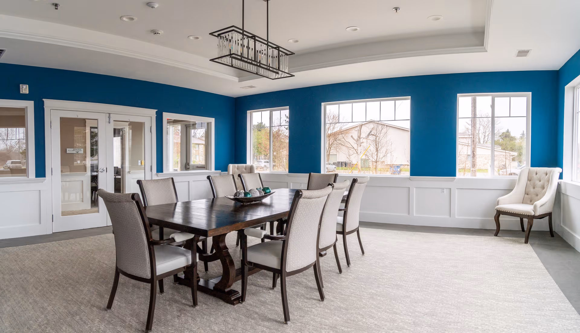 Bright dining room with a long wooden table surrounded by upholstered chairs, blue accent walls, and large windows.