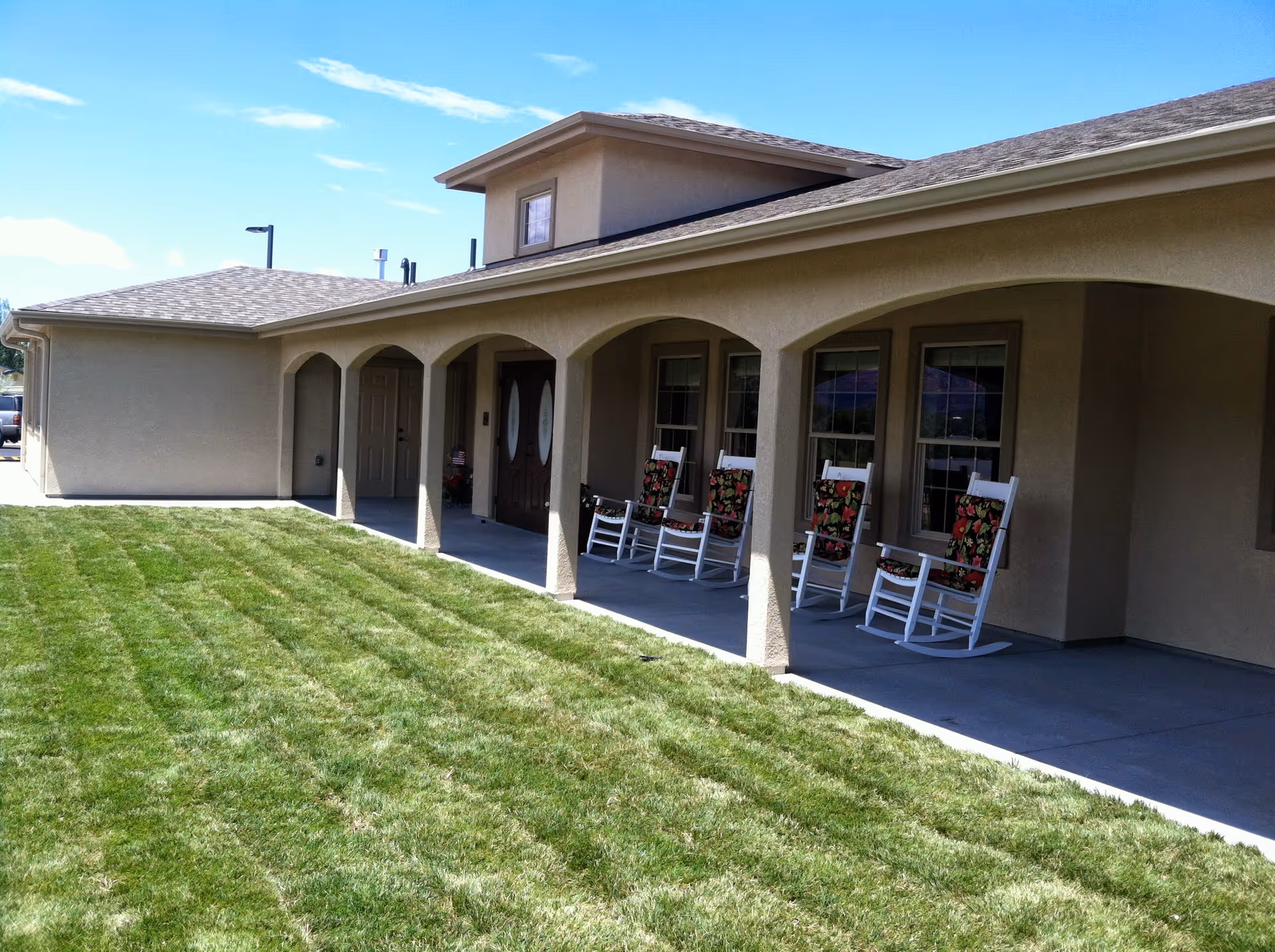 Exterior view of a single-story assisted living facility building with a covered porch featuring several white rocking chairs with floral cushions. The building has beige walls, arched openings, and a well-maintained green lawn in front under a partly cloudy blue sky.