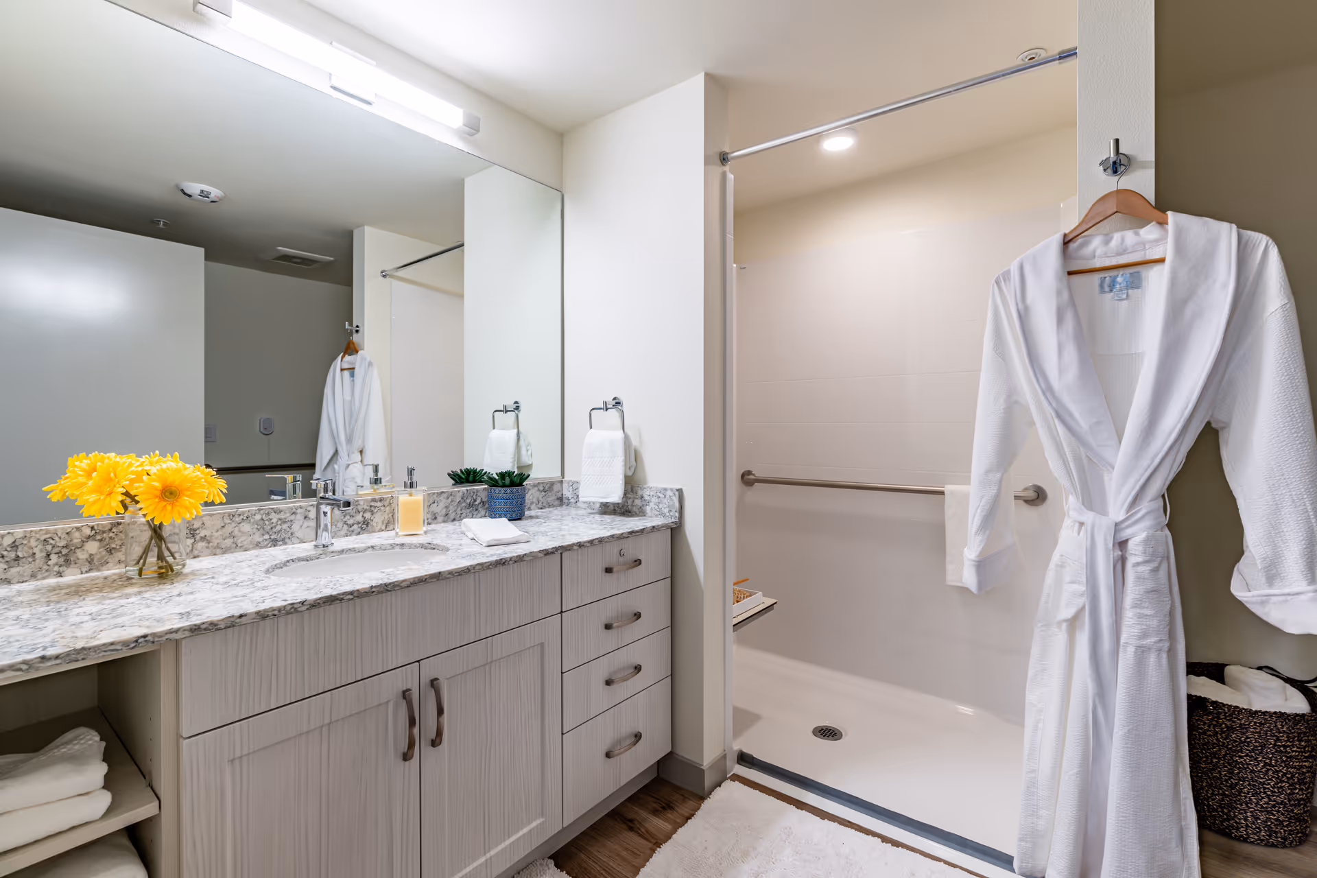 Well-lit bathroom with a marble countertop vanity, large mirror, open walk-in shower and a white bathrobe hanging on a hook.