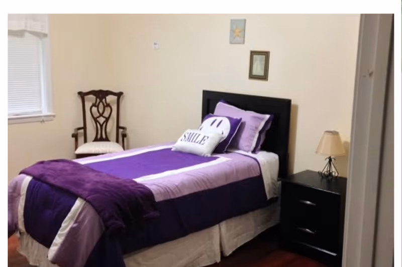 Small bedroom with a single bed dressed in purple bedding, a nightstand with a lamp, and a wooden chair by the window.