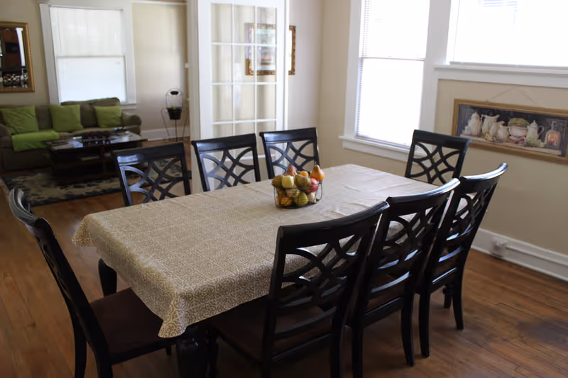 A dining room with a rectangular table covered with a beige patterned tablecloth and surrounded by eight dark wooden chairs with decorative backs. A fruit basket centerpiece sits on the table. In the background, there is a living room area with a green couch, a coffee table, and a rug. The room has wooden floors, white walls, and windows with blinds allowing natural light to enter.