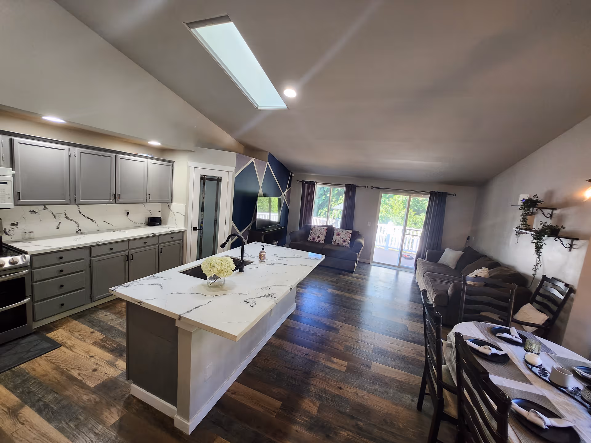 Open-plan kitchen with a large marble island opening to a living room with sofas and sliding glass doors.