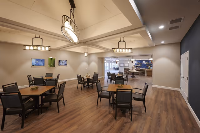 Open dining room with several square wooden tables and chairs, pendant lighting, and wood-look flooring leading to a serving area.