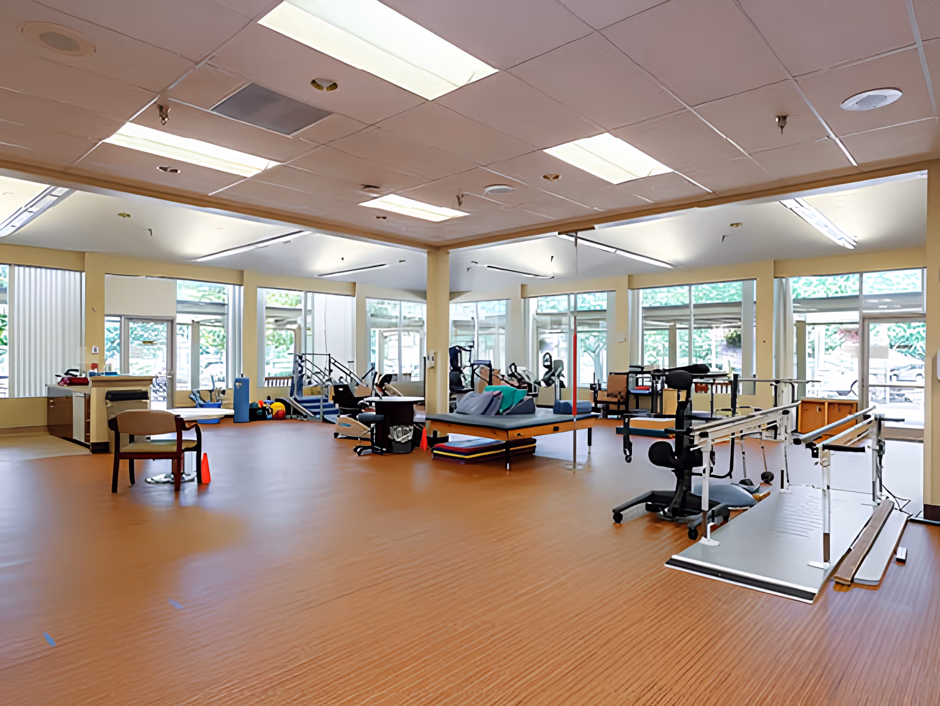 A spacious physical therapy or rehabilitation room with exercise equipment, parallel bars, therapy tables, chairs, and large windows letting in natural light.