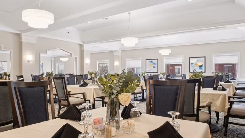 A bright and elegant dining room with multiple tables covered in beige tablecloths, each set with black napkins, upside-down wine glasses, and a centerpiece of green foliage and white flowers. The room features large windows, framed abstract artwork on the walls, and modern ceiling lights.