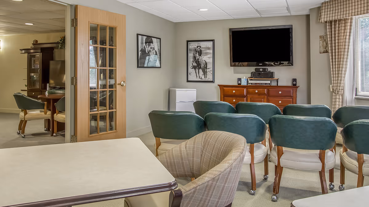 A cozy common area in a senior living community featuring a row of green cushioned chairs facing a wall-mounted flat screen TV above a wooden cabinet. There is a small white refrigerator next to the cabinet, framed black and white pictures on the wall, and a window with plaid curtains letting in natural light. A plaid armchair and tables are also visible, with a glass-paneled wooden door leading to another room with additional seating and a TV.