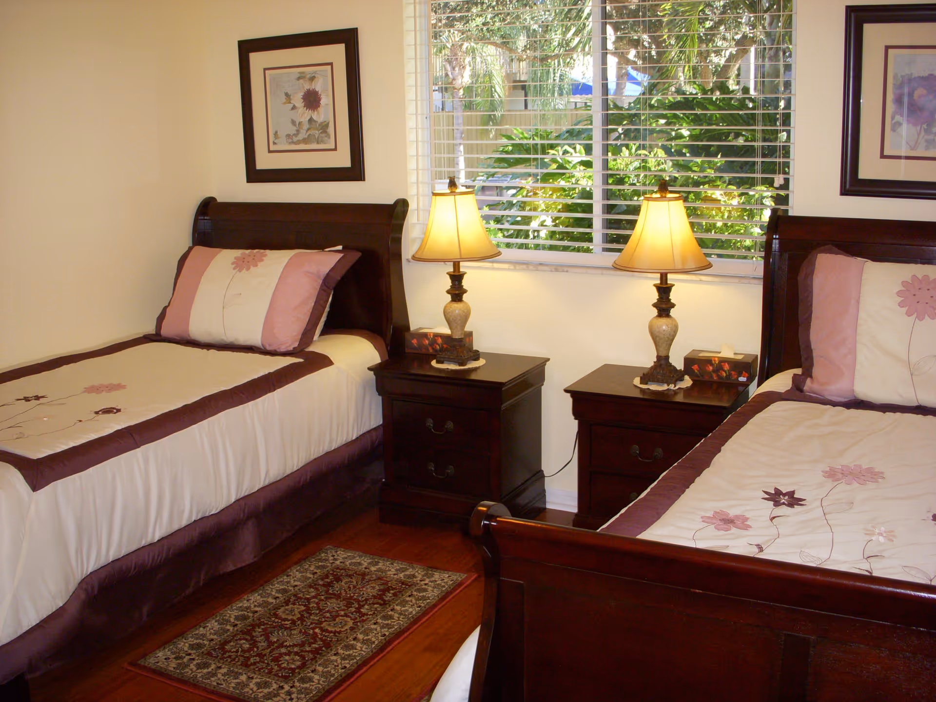 Twin bedroom with two wooden beds, matching nightstands and lamps beneath a window with blinds and greenery outside.
