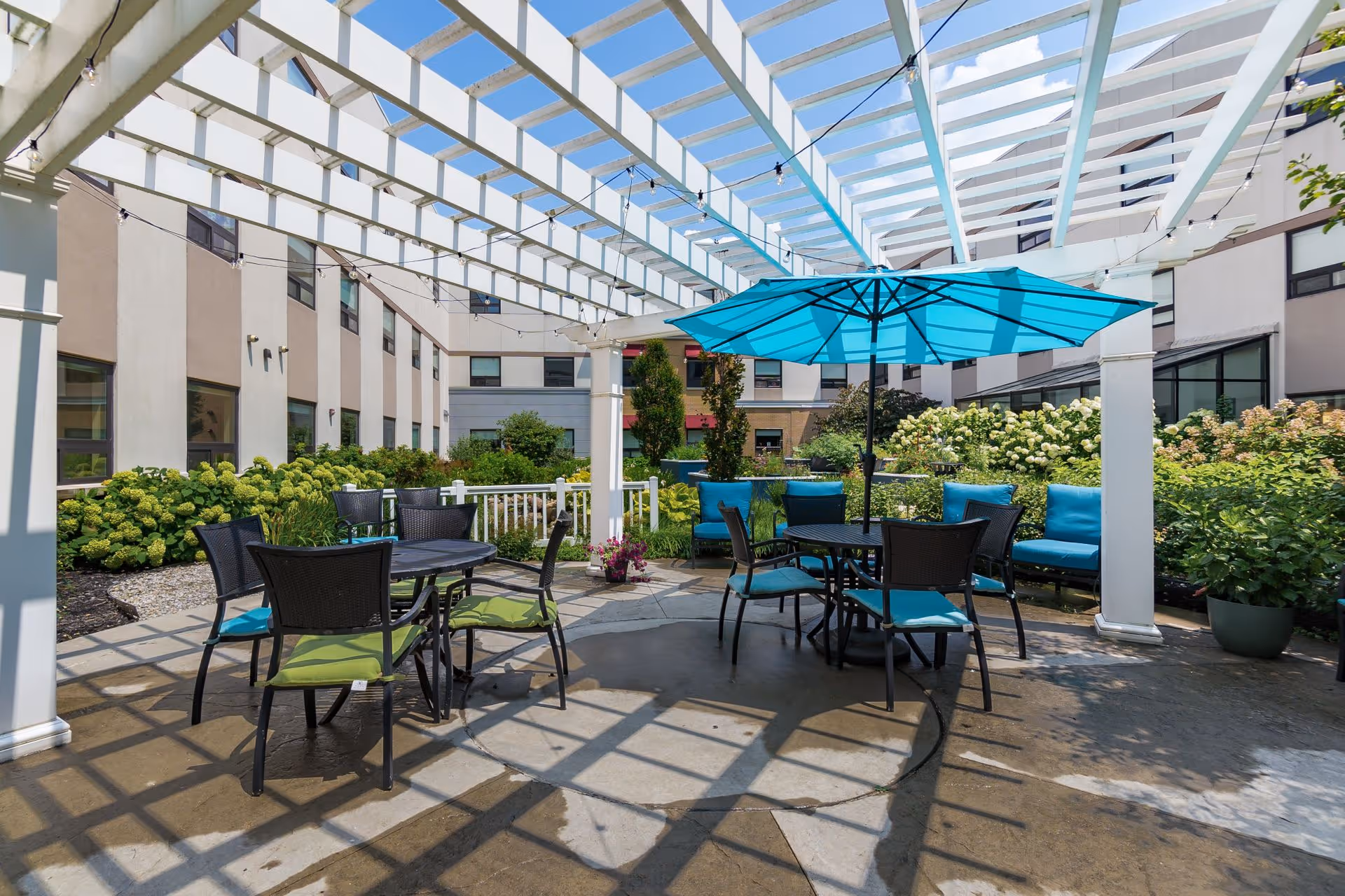 Outdoor patio area at The Avalon of Lewis Center featuring a white pergola overhead, two round tables with chairs, one table shaded by a large blue umbrella, surrounded by green plants and flowers with the building in the background.