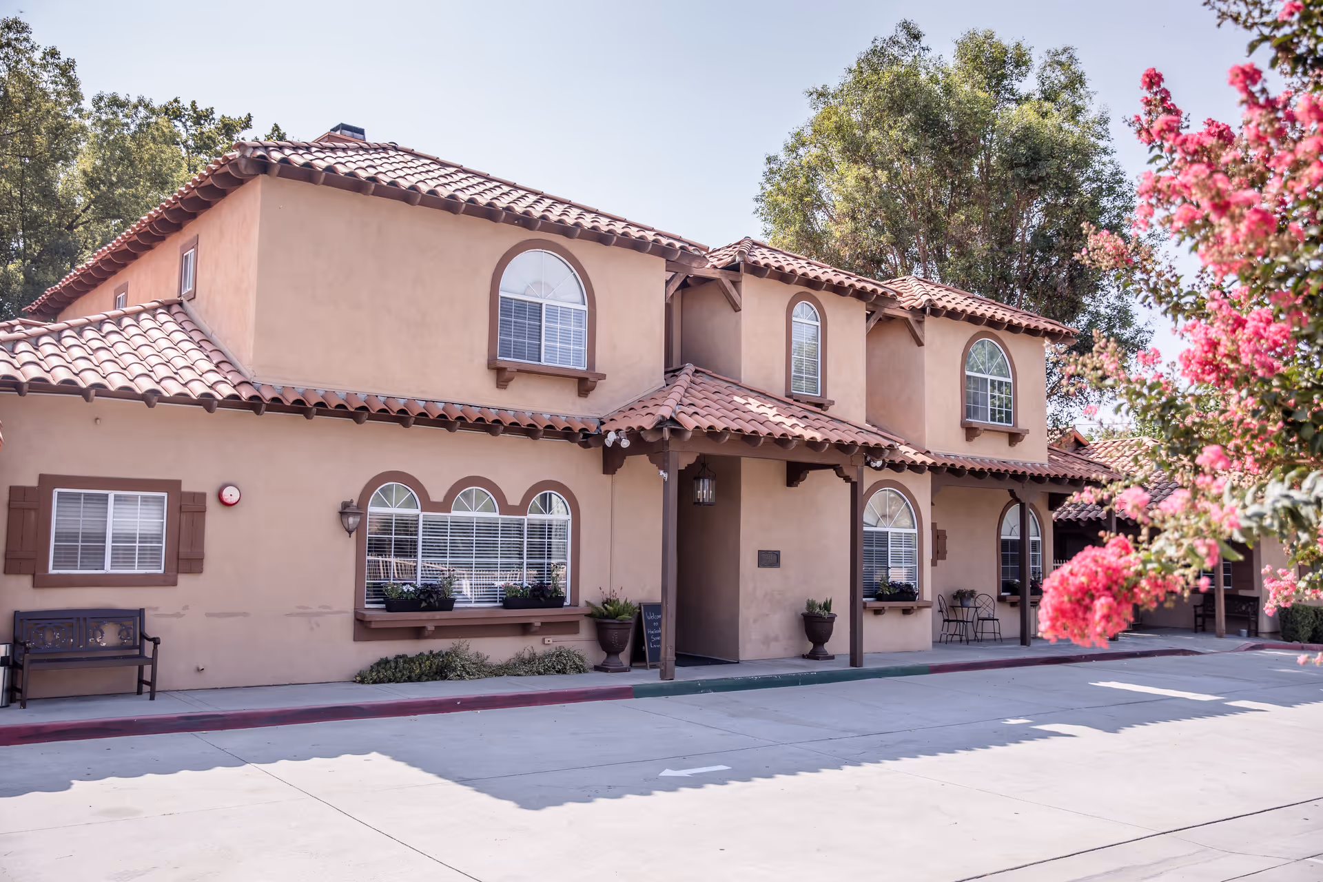 Exterior view of a two-story building with a terracotta tiled roof and beige stucco walls. The building has multiple arched windows with white blinds and brown shutters. There are potted plants near the entrance and a bench on the left side. Pink flowering trees are visible on the right side of the image under a clear sky.