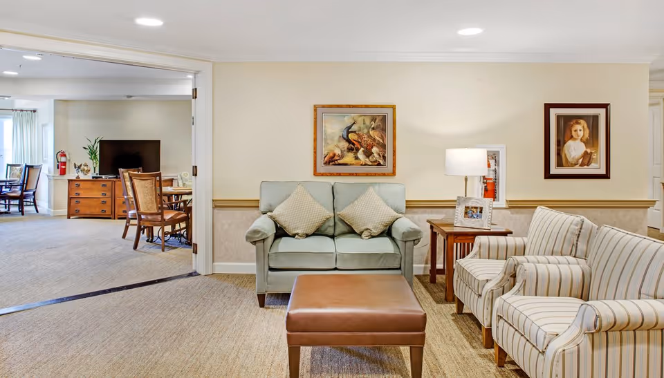 Comfortable common living room with a sofa, armchairs, ottoman, side table and framed artwork, with a dining area visible through an open doorway.