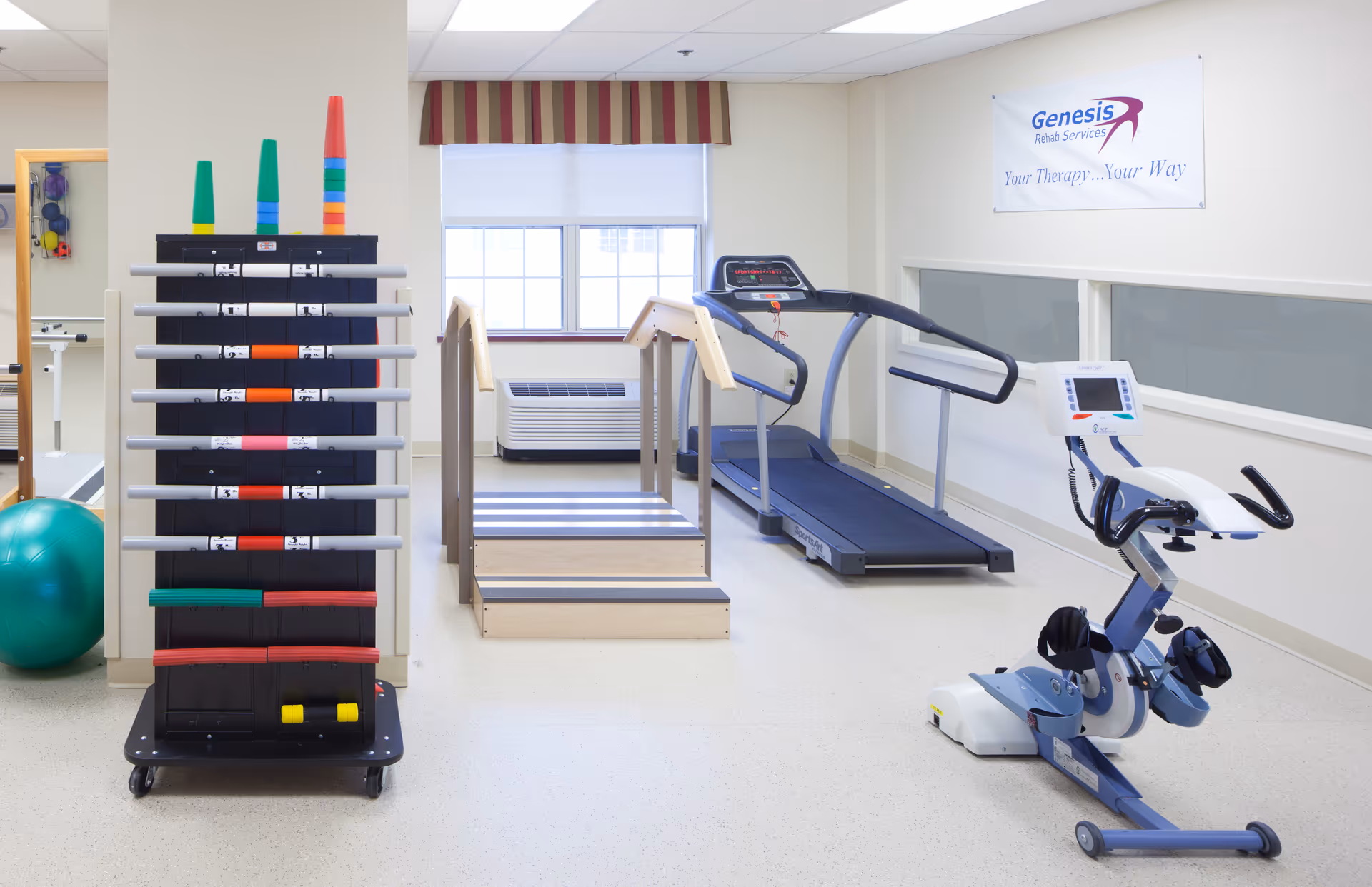 A rehabilitation therapy room with exercise equipment including a treadmill, a stationary bike, a set of stairs with handrails, and a rack holding various colored exercise bars. A green exercise ball is partially visible on the left. A window with a striped valance lets in natural light, and a sign on the wall reads 'Genesis Rehab Services Your Therapy... Your Way'.