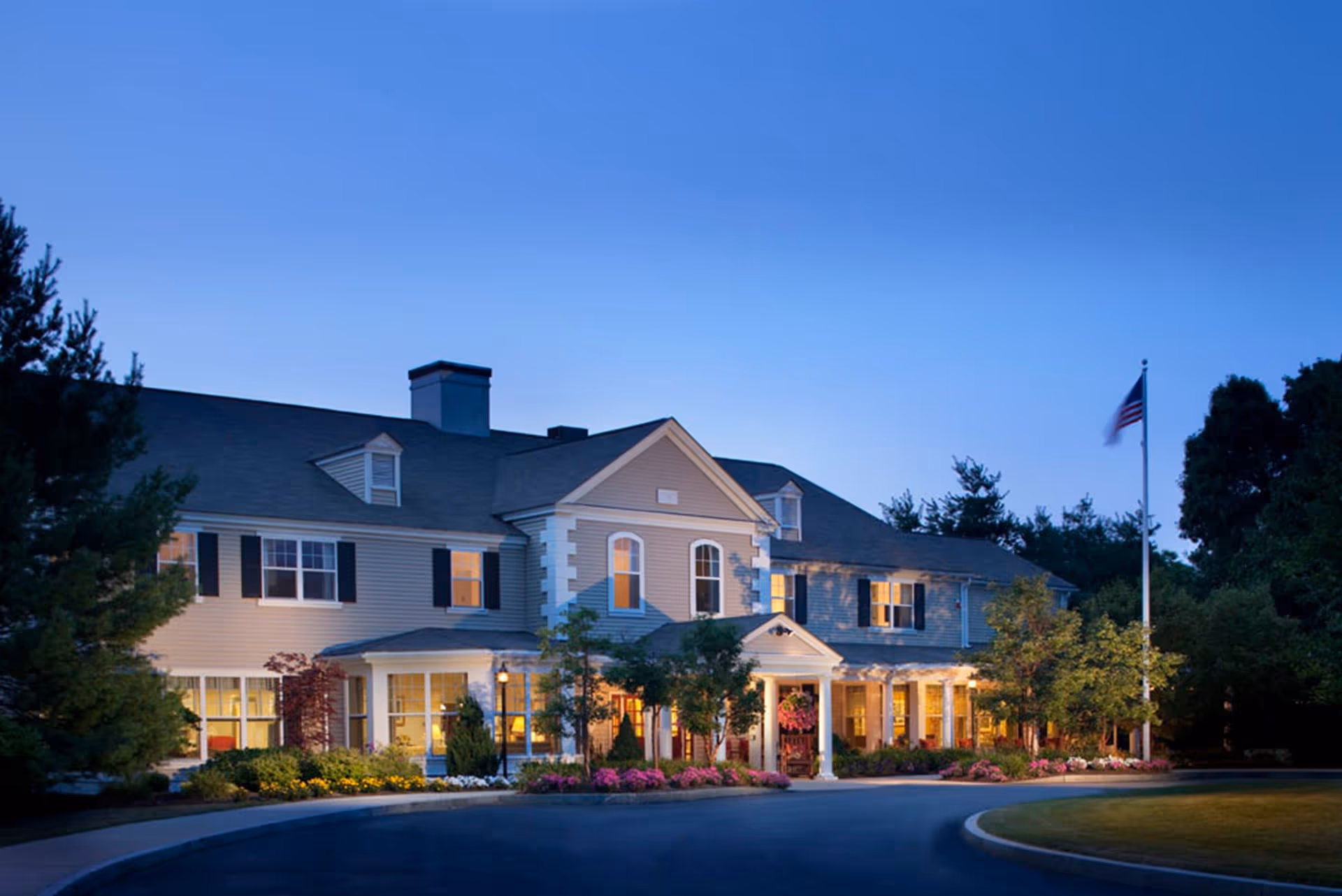 Exterior view of a large two-story residential building at dusk with lights glowing from the windows. The building has a pitched roof, multiple windows with shutters, and a covered entrance with columns. There are landscaped bushes, flowers, and trees around the building, and an American flag on a flagpole to the right.