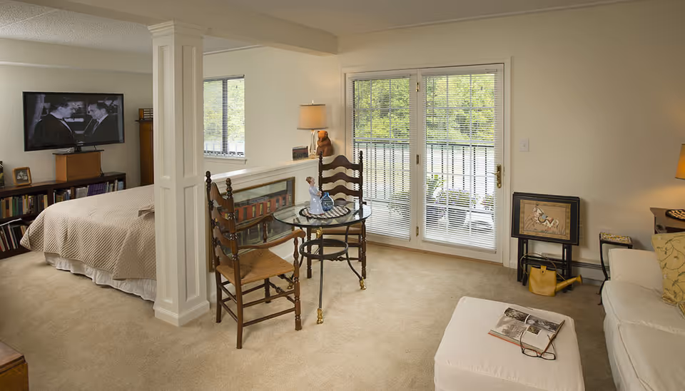 Bright studio-style interior showing a bed, TV, bookshelf, small glass dining table with chairs, sofa and ottoman, and glass doors to a balcony.