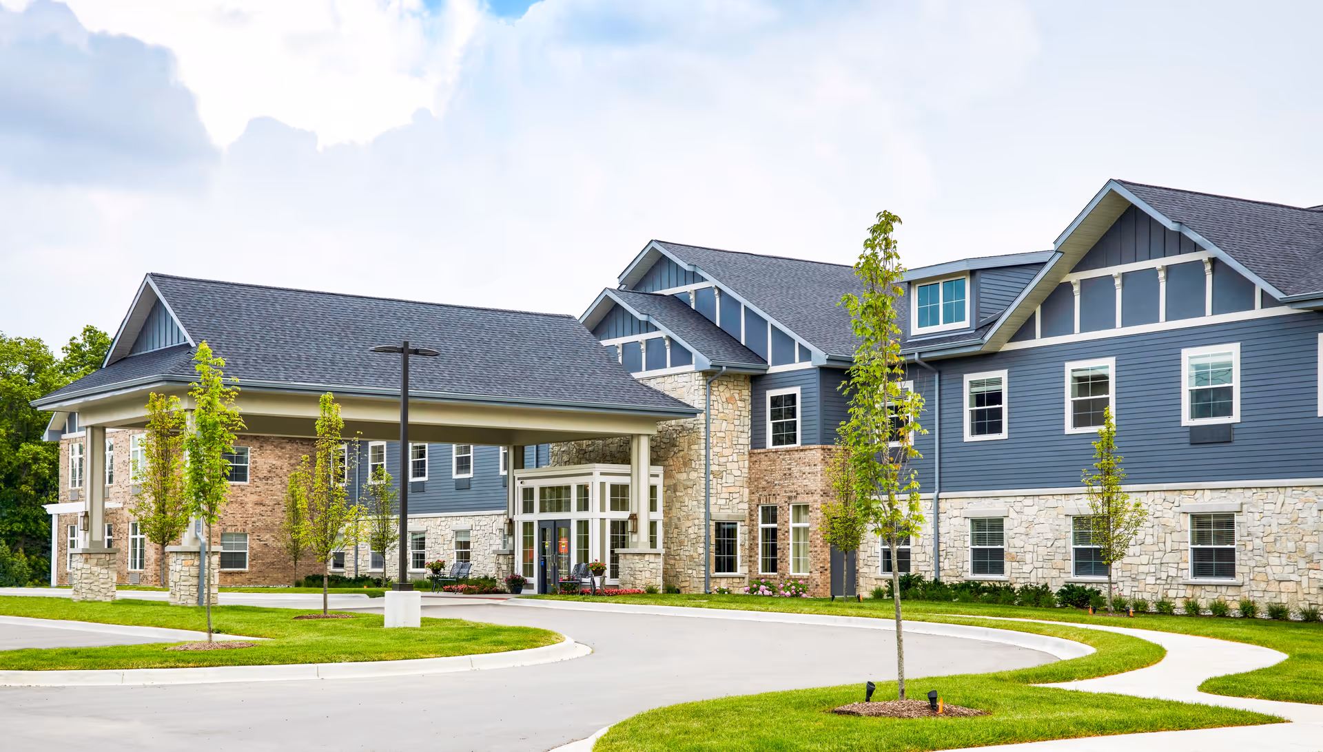 Exterior view of a modern senior living facility building with a covered entrance, multiple windows, and a combination of stone and blue siding. The surrounding area features a curved driveway, green grass, young trees, and a partly cloudy sky.