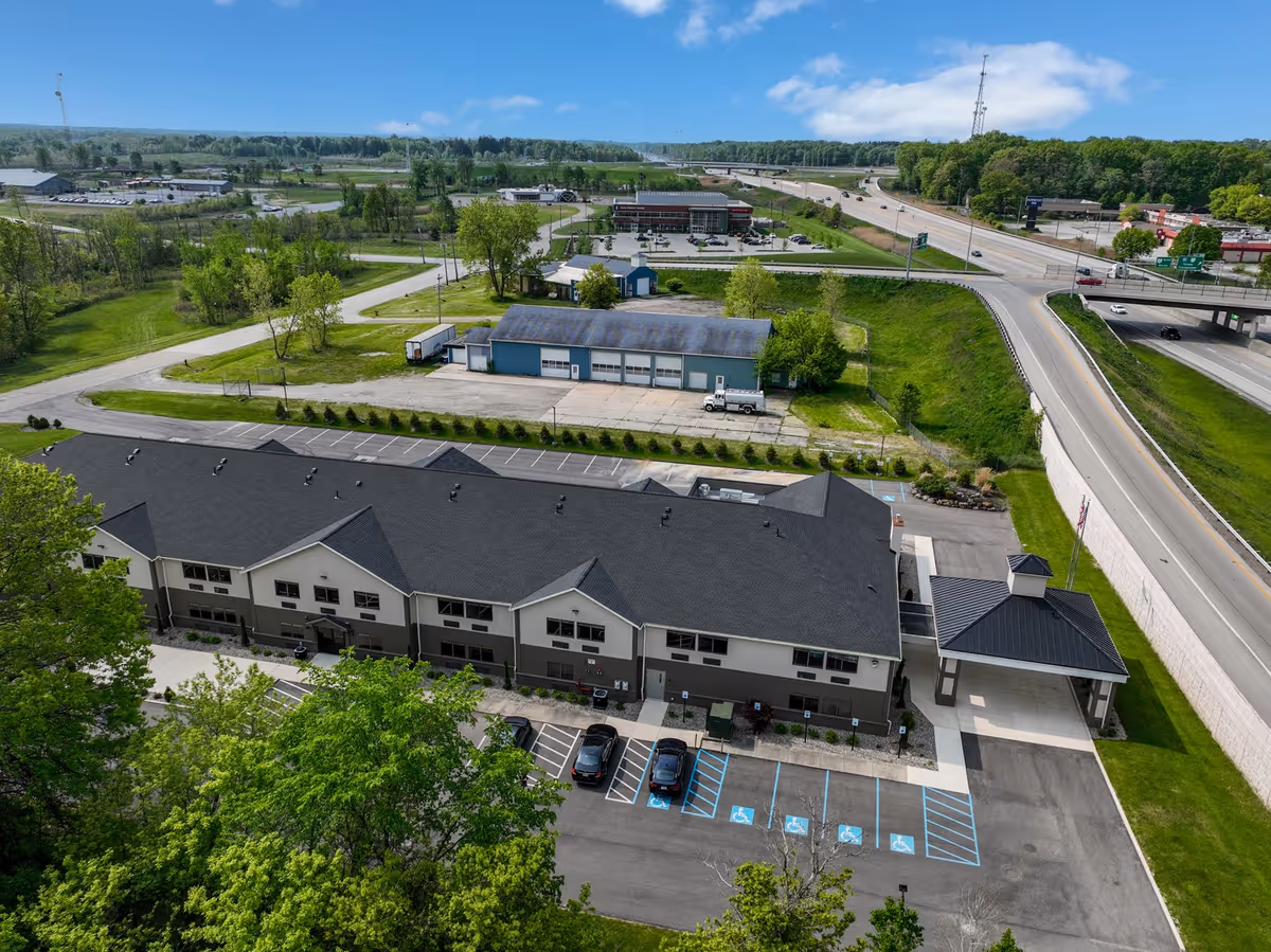 Aerial view of Hines Hill Senior Living facility showing a two-story building with a dark roof, surrounded by parking spaces including several handicapped spots. The building is adjacent to a highway and surrounded by green trees and grassy areas. Other commercial buildings and roads are visible in the background under a clear blue sky.