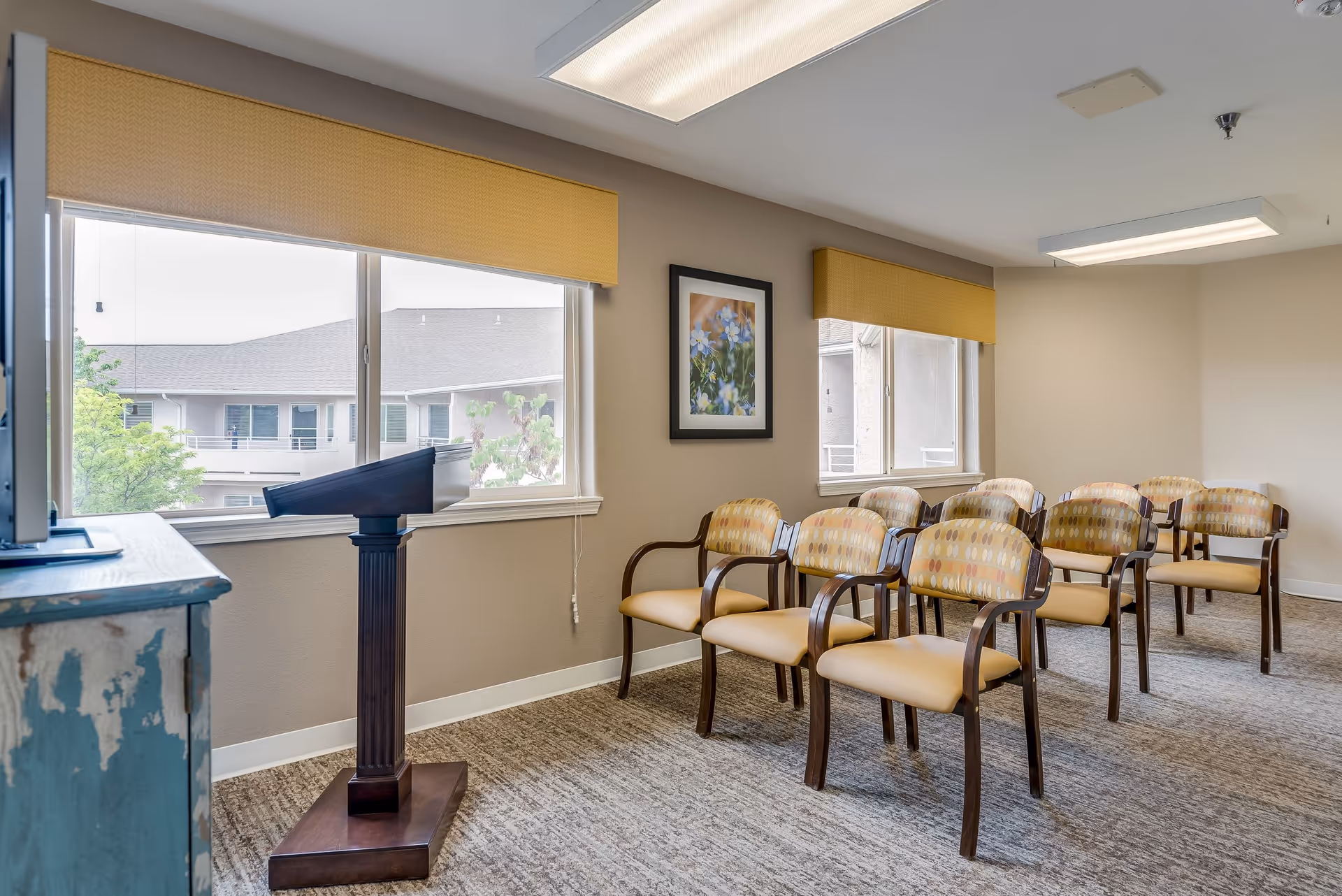 A small conference or meeting room with rows of wooden chairs featuring patterned cushions. There is a wooden podium at the front near large windows with yellow valances, and a framed picture of flowers on the beige wall. The room is carpeted and has fluorescent ceiling lights.