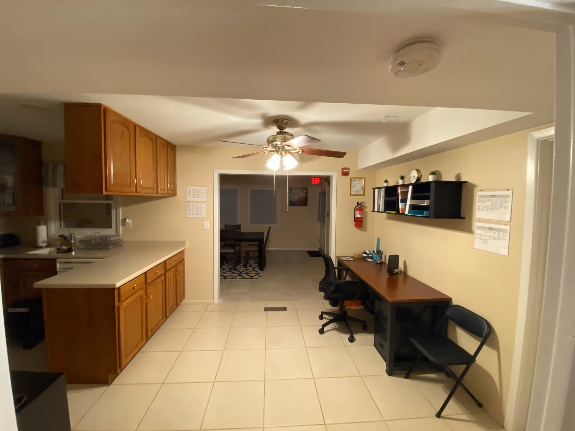 Interior view of a room in Holy Hills Assisted Living featuring a kitchen area with wooden cabinets and a countertop on the left, and a workspace on the right with a desk, office chair, folding chair, and wall-mounted shelves holding office supplies. A ceiling fan with lights is in the center of the ceiling, and an adjoining room with a dining table and chairs is visible through an open doorway.