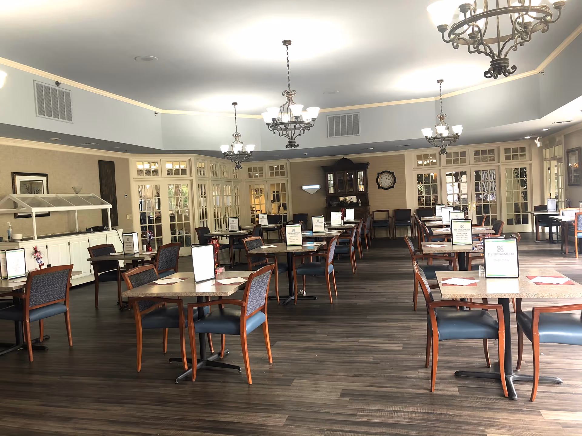 A spacious dining room with multiple square tables and chairs arranged neatly. Each table has a menu or sign placed on it. The room features chandeliers hanging from the ceiling, large windows, and glass-paneled doors along the walls. The floor is dark wood, and there is a buffet counter on the left side of the room.