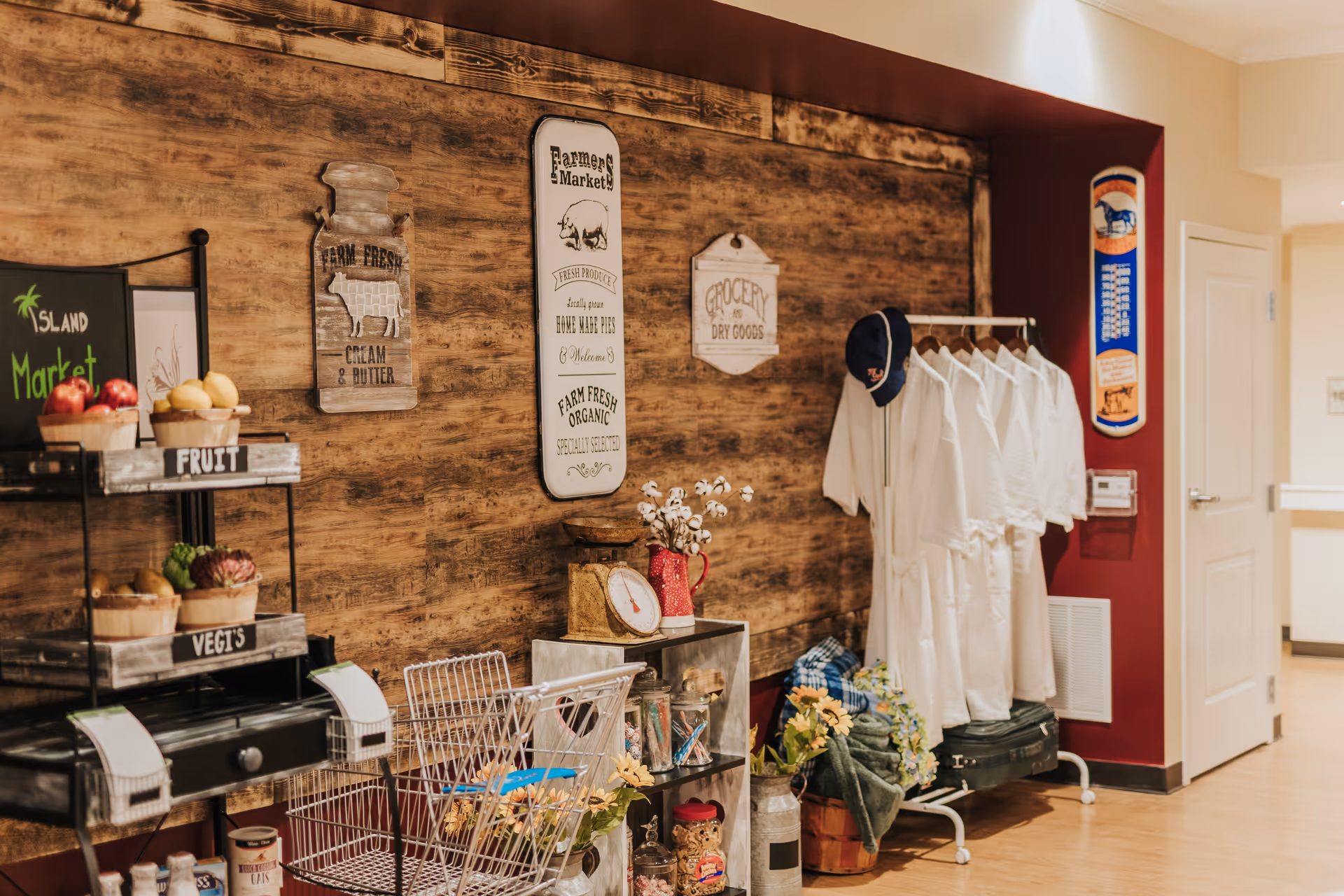 Interior view of a room with a rustic wooden wall decorated with vintage-style signs about farm fresh products. There is a black metal shelf labeled 'Island Market' holding baskets of fruit and vegetables. Nearby, a small metal cart holds jars and a scale, and a clothing rack displays several white robes and a blue cap. The floor is wooden, and the walls are painted beige and dark red.