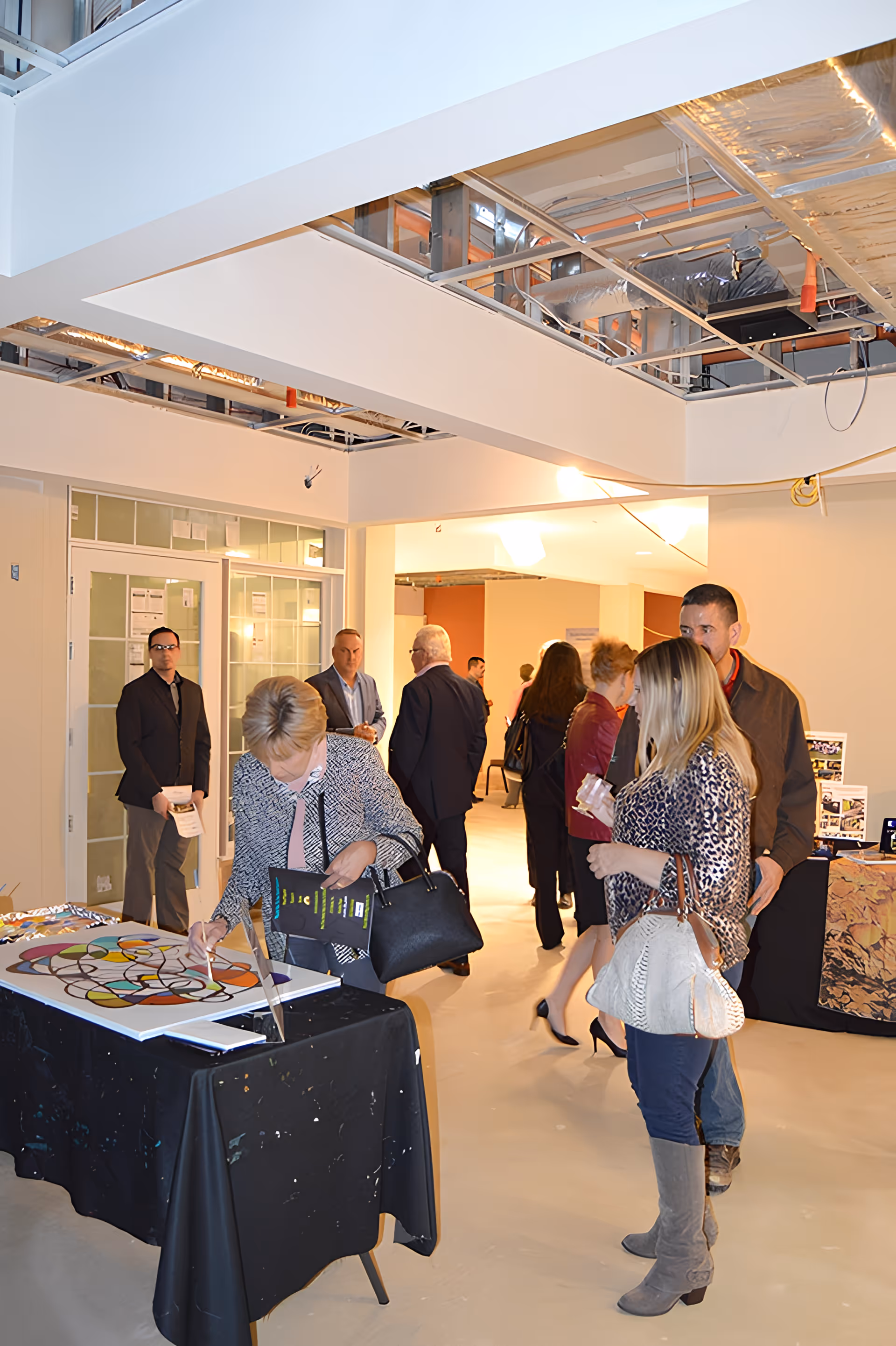 A group of people gathered indoors at an event in a building under construction or renovation. Some individuals are looking at artwork displayed on tables covered with black cloths. The ceiling is partially open, exposing ducts and wiring. The atmosphere appears to be a casual social or networking event.