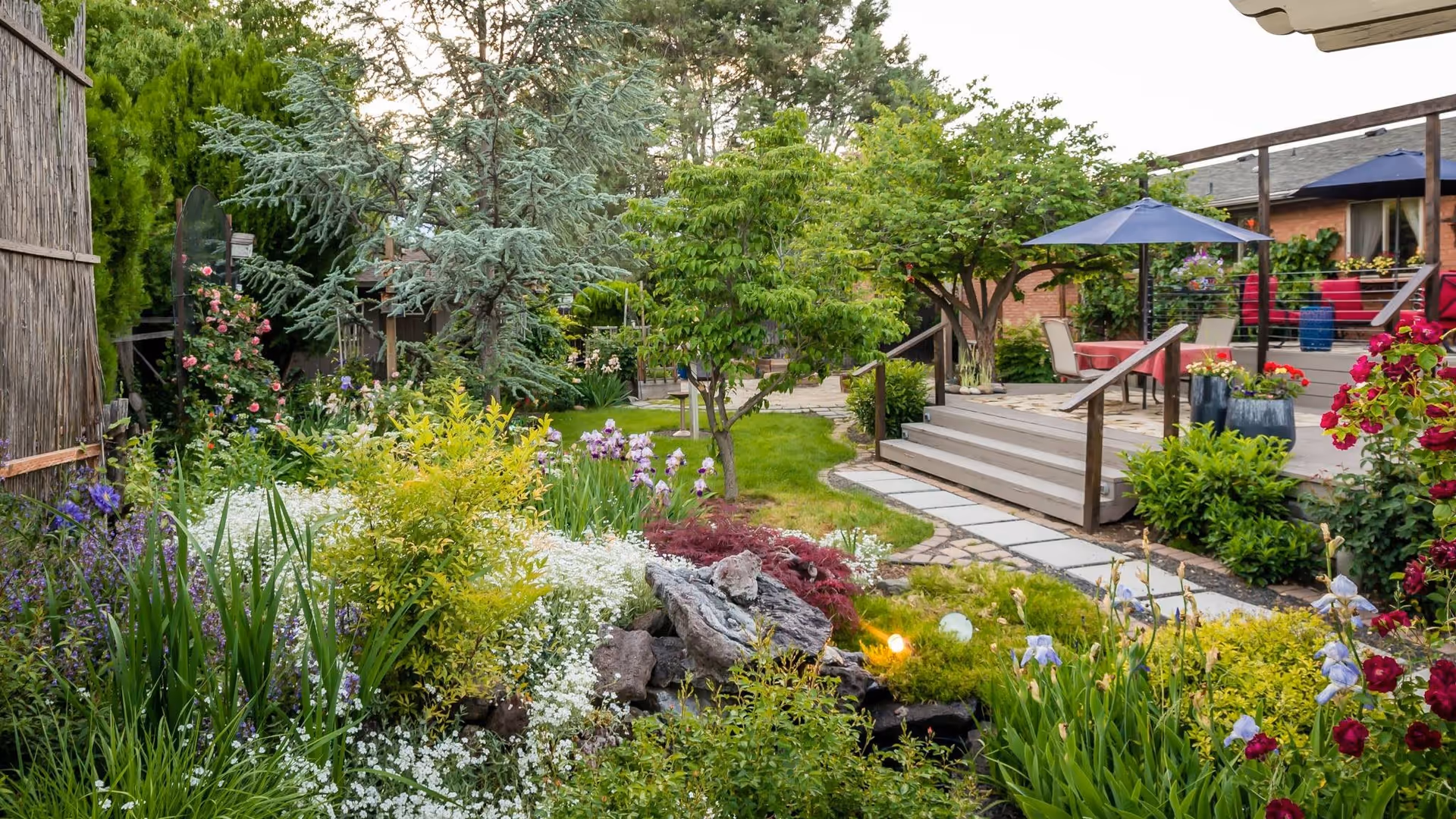 A lush garden area with various green plants, colorful flowers, and small trees. A stone pathway leads to a wooden deck with outdoor seating, tables, and blue umbrellas. The setting appears peaceful and well-maintained.