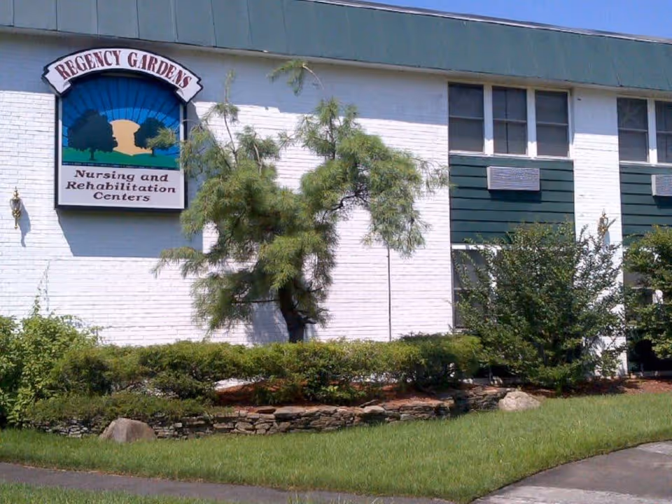 Exterior view of a building with white brick walls and green paneling. There is a sign on the building that reads 'Regency Gardens Nursing and Rehabilitation Centers'. The foreground features a landscaped area with bushes, a small tree, rocks, and a grassy lawn.