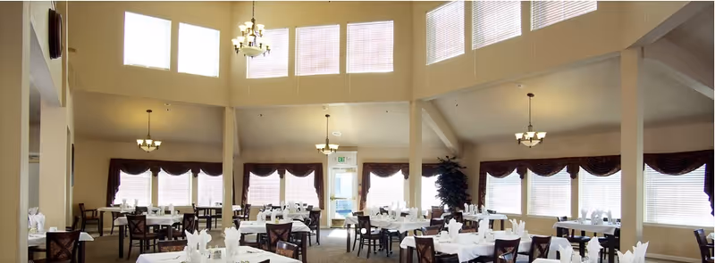 A spacious dining room with multiple tables covered in white tablecloths and neatly arranged napkins. The room features high ceilings with several chandeliers and large windows with blinds and dark valances, allowing natural light to fill the space.
