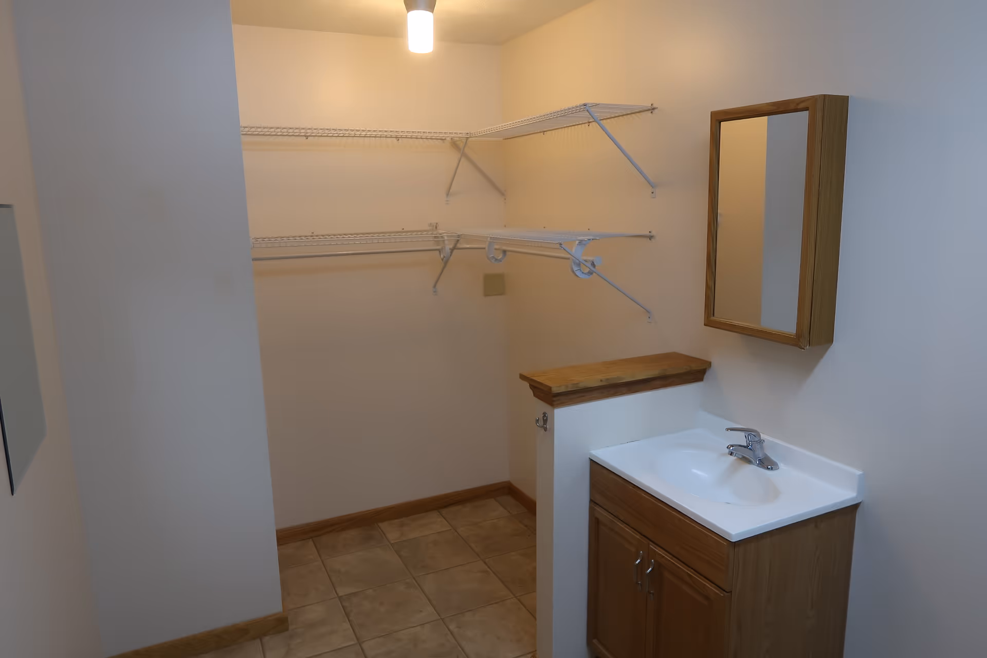 Small interior room with a sink and vanity, wall-mounted mirror cabinet, tile floor, ceiling light, and wire shelving.