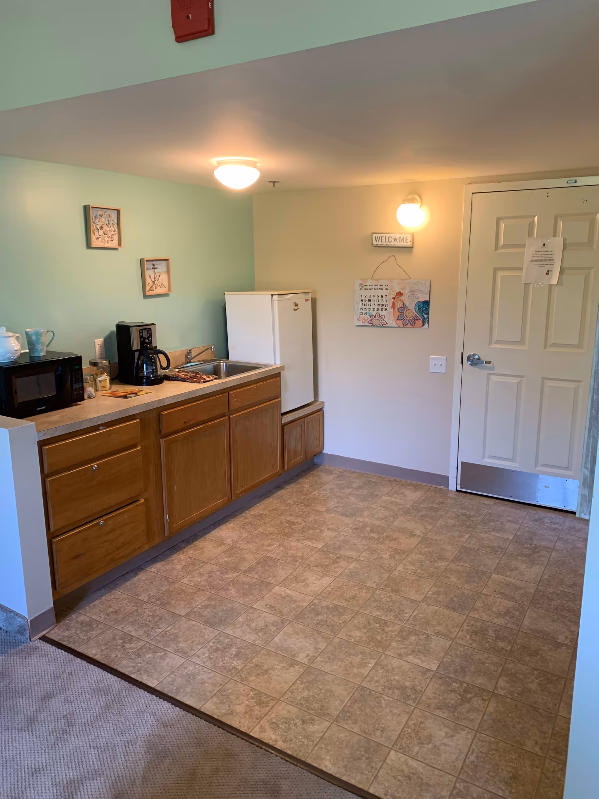 A small kitchenette area with wooden cabinets, a countertop with a microwave, coffee maker, and sink. There is a white mini refrigerator next to the cabinets. The floor is tiled, and the walls are painted light green and beige. A door is visible on the right side with a calendar and a small 'WELCOME' sign hanging on the wall above a light fixture.