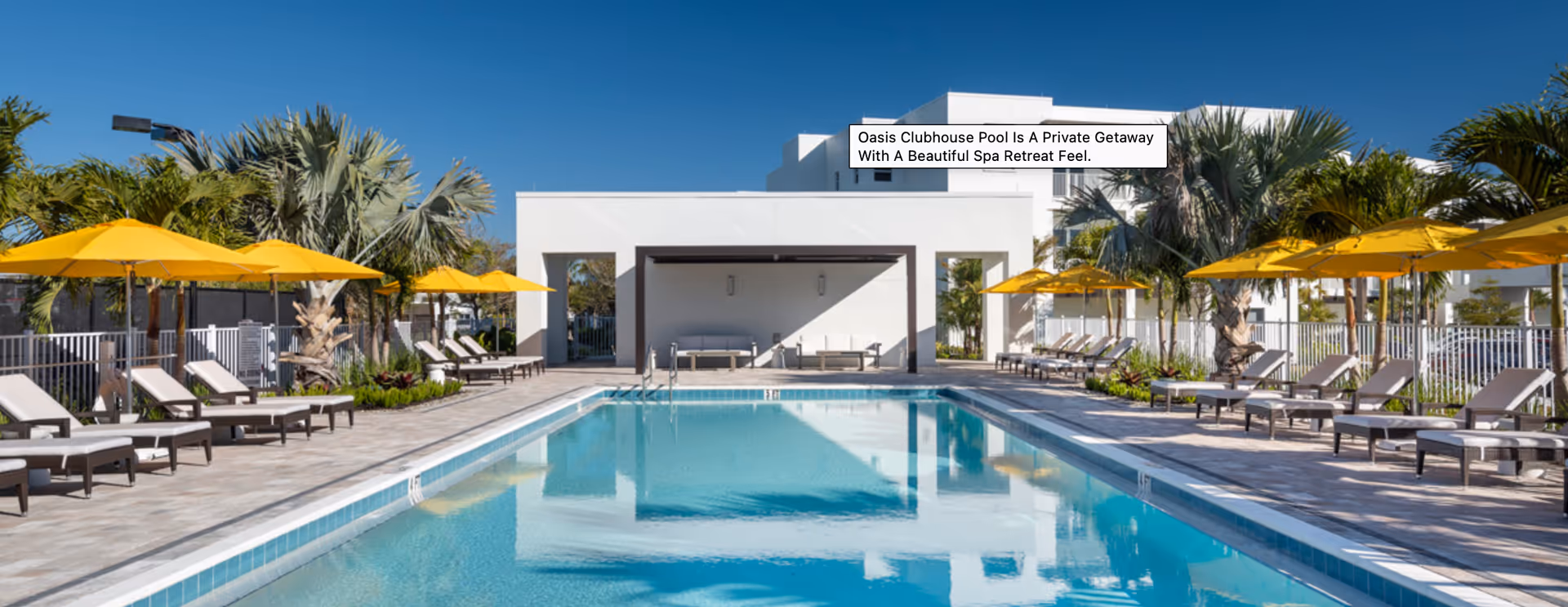 Outdoor swimming pool area at a senior living facility with lounge chairs and yellow umbrellas on both sides, palm trees, and a white clubhouse building in the background under a clear blue sky.