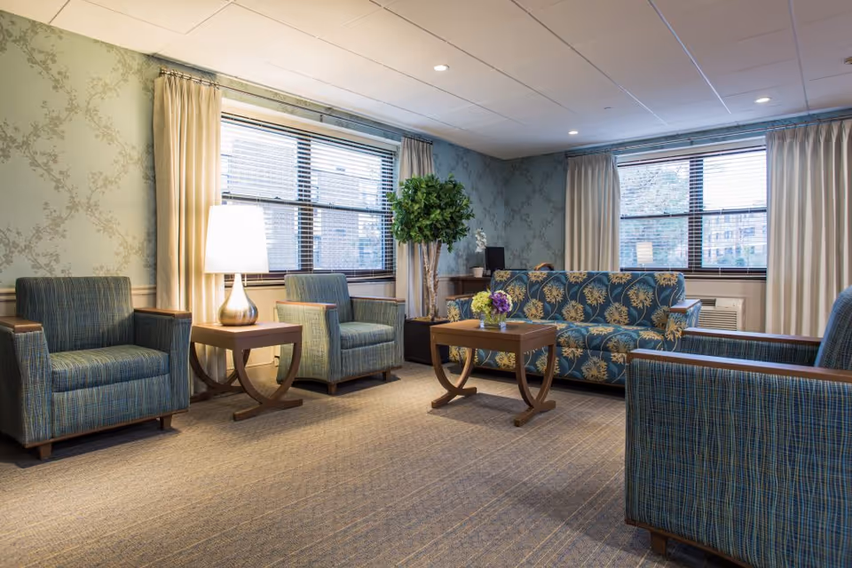 A cozy living room area with patterned wallpaper, two large windows with beige curtains, a blue floral sofa, three blue armchairs with wooden armrests, a wooden coffee table with a small flower arrangement, a side table with a lamp, and a potted plant in the corner.