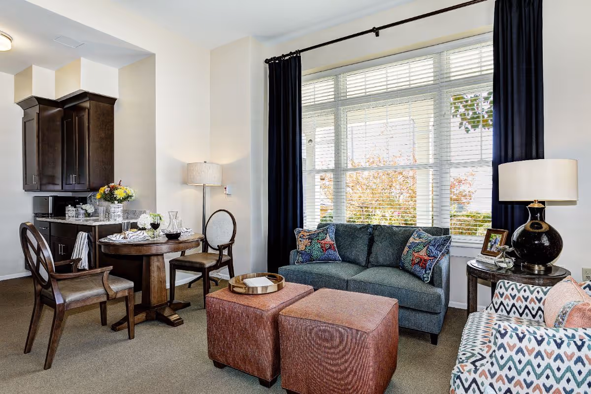 A cozy living room area with a large window covered by blinds and dark curtains. The room features a blue sofa with decorative pillows, a patterned armchair, two square ottomans with a tray on top, a round wooden side table with a lamp and picture frame, and a small round dining table with two chairs. In the background, there is a kitchen area with dark wooden cabinets and a countertop with flowers and glassware.
