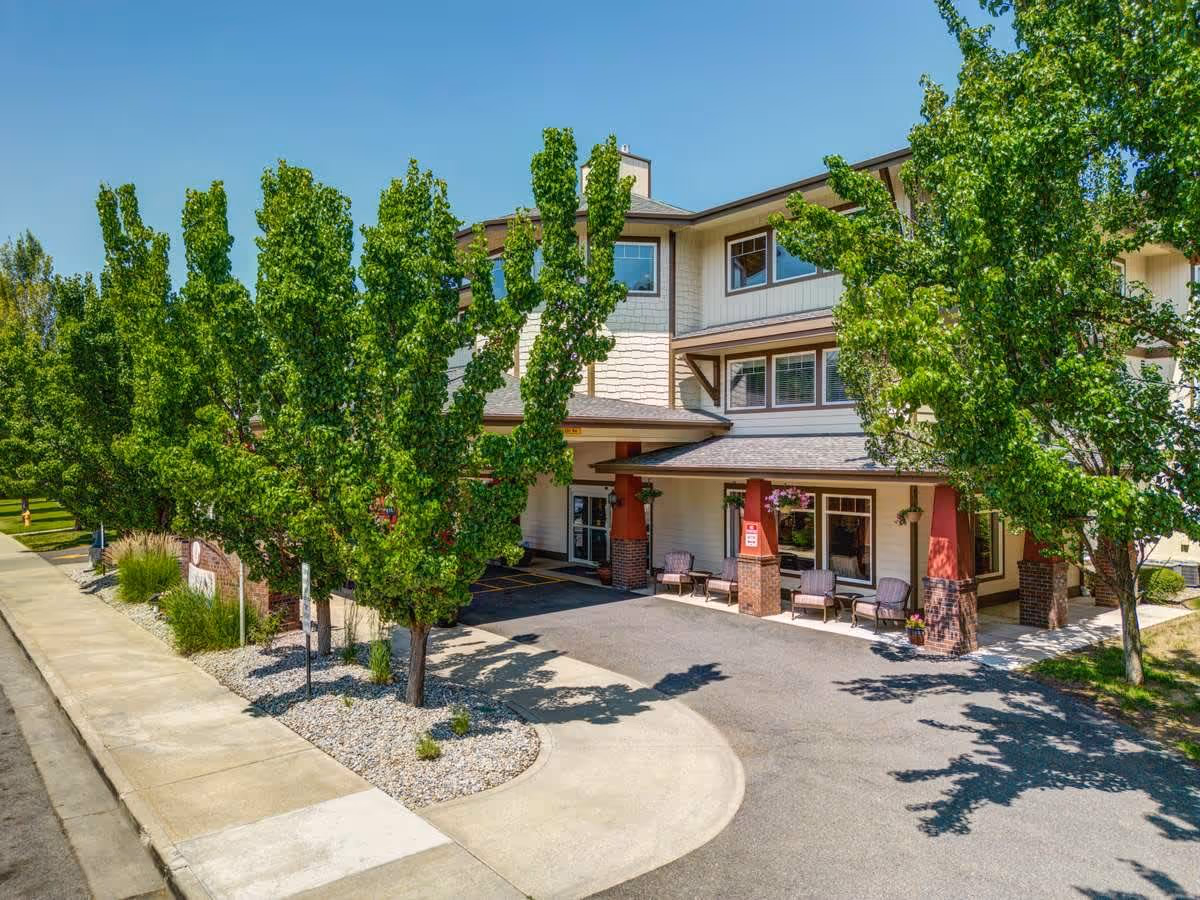 Exterior view of a multi-story senior living facility named South Hill Village, featuring a covered entrance with seating and surrounded by green trees and landscaping under a clear blue sky.