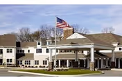 Front exterior of a multi-story senior living facility with a covered porte-cochere and an American flag.