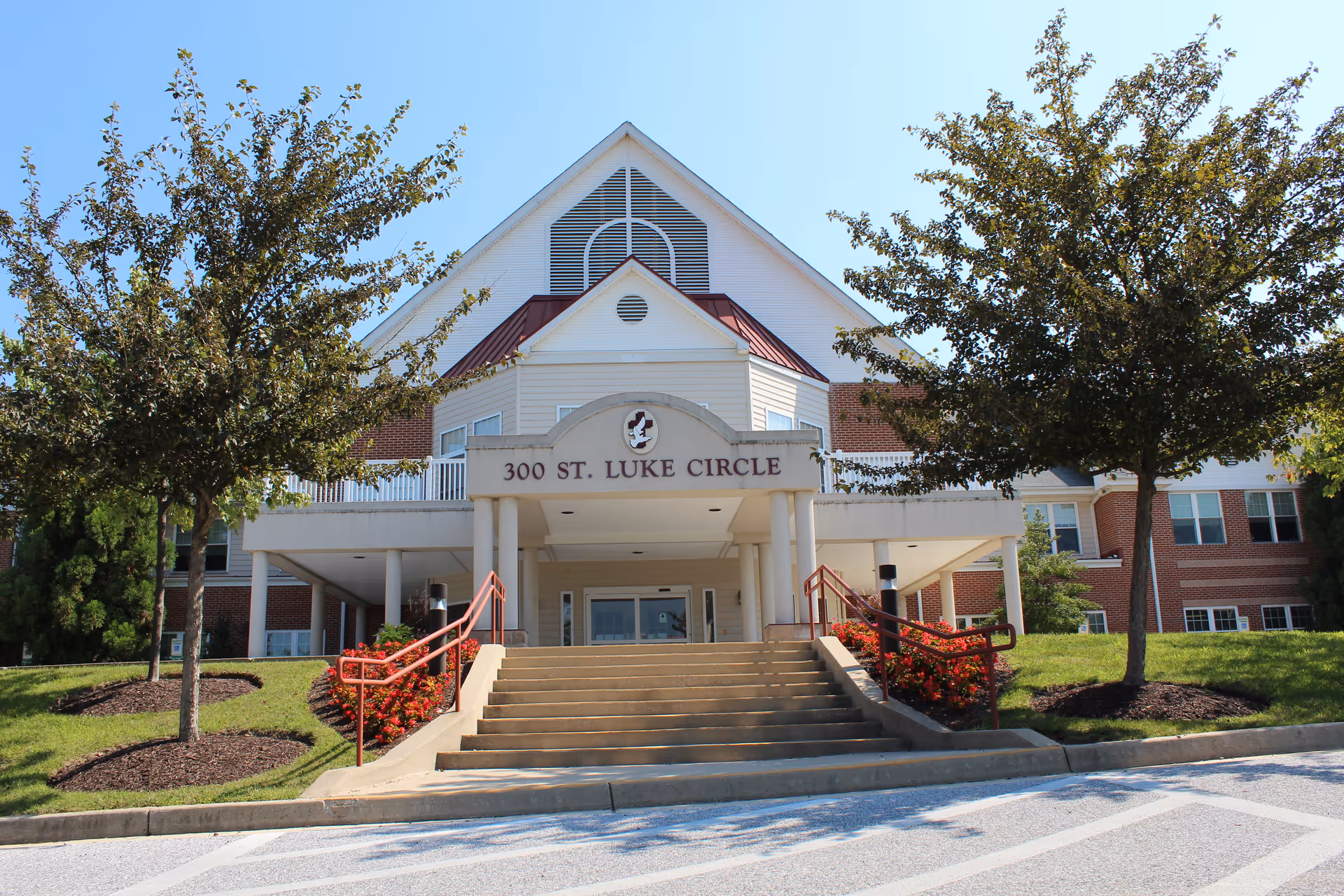 Front exterior view of a senior living facility building with a sign reading '300 St. Luke Circle'. The building has a peaked roof, red railings along the stairs leading to the entrance, and is flanked by two trees with green foliage. The sky is clear and blue.