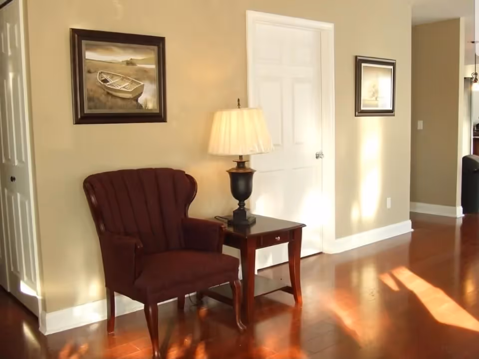 A cozy corner of a room with a maroon upholstered armchair next to a wooden side table holding a lamp with a beige shade. The walls are painted light beige and decorated with two framed paintings. The floor is polished wood, and sunlight casts shadows across it.