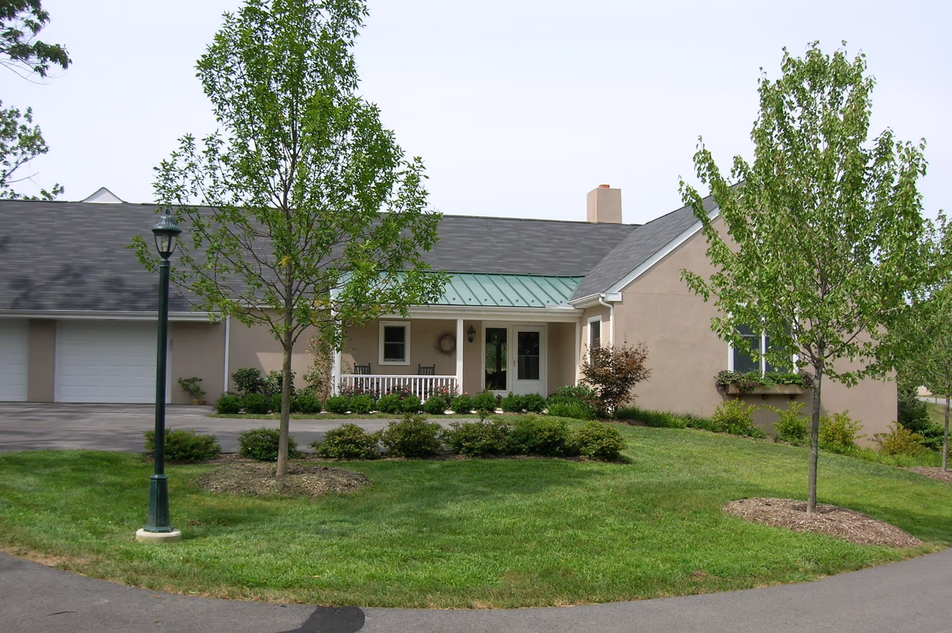 Exterior view of a single-story building with a gray roof and beige walls, featuring a small covered porch with two chairs. The building is surrounded by green grass, small bushes, and young trees, with a paved driveway and a streetlamp in front.