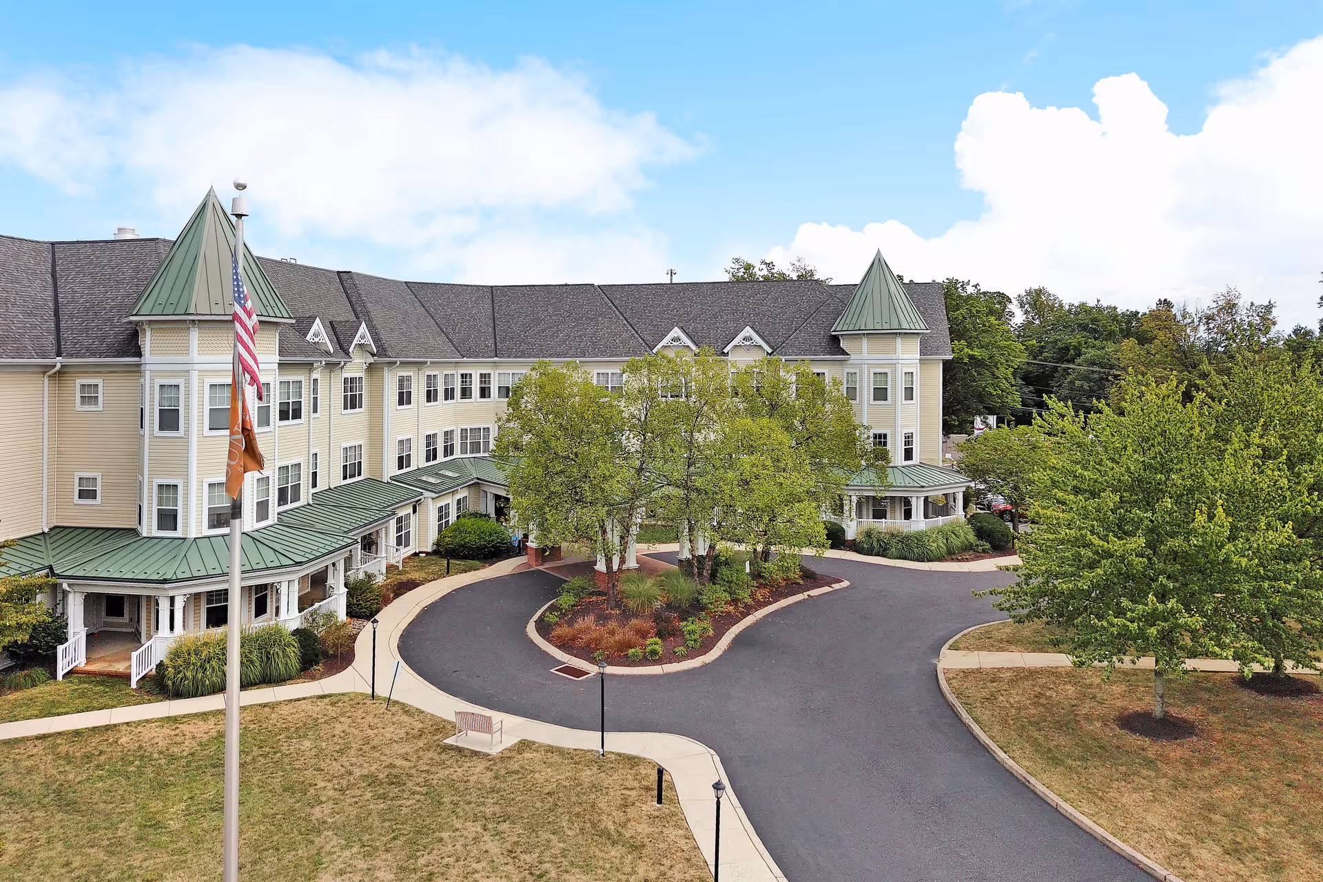 Exterior view of a large senior living facility building with beige siding and green roofs, featuring two turret-like structures. There is a circular driveway with landscaped trees and bushes in the center, an American flag and another flag on a flagpole in the foreground, and a partly cloudy sky above.
