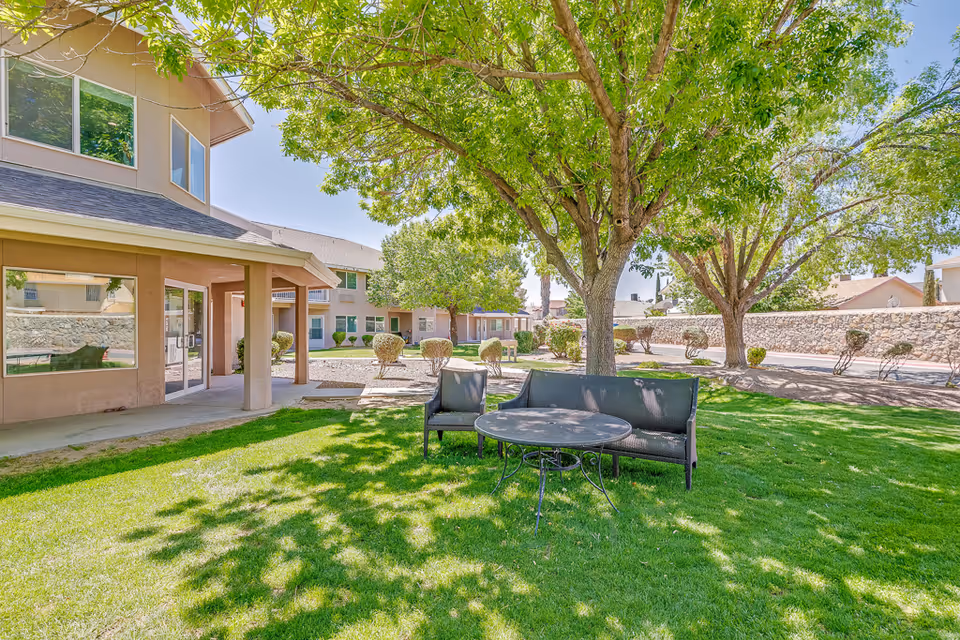 Outdoor seating area with a black metal table and two black cushioned chairs on a green lawn under large leafy trees. In the background, there are beige buildings with large windows and a stone wall surrounding the property under a clear blue sky.