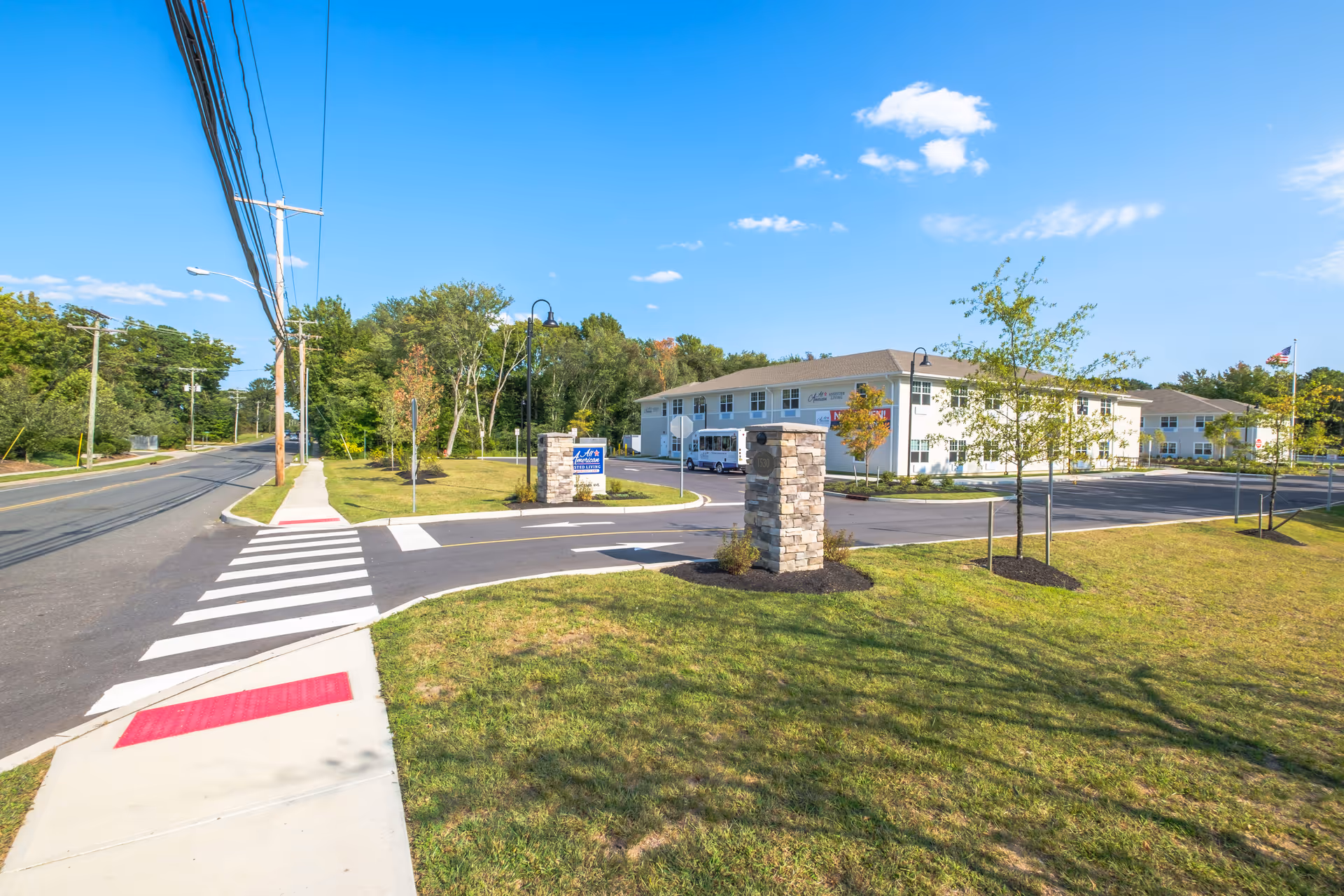 Exterior view of All American Assisted Living at Tinton Falls facility on a sunny day, showing a two-story building with a parking lot, landscaped lawn with young trees, a stone pillar with the address 1530, and a sign for the facility near the entrance. A sidewalk and crosswalk are visible in the foreground along a quiet street with utility poles and trees lining the road.