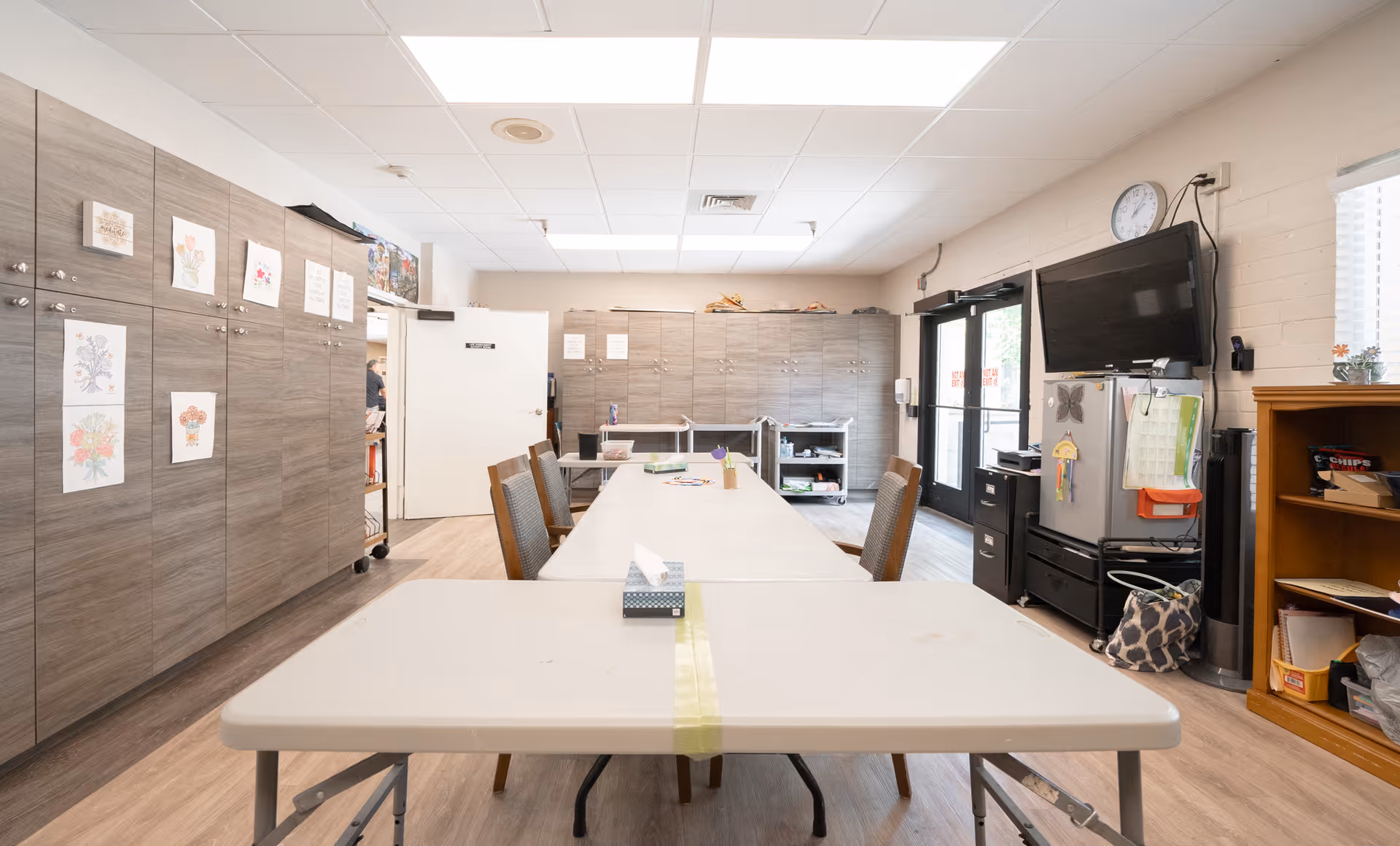 A bright room with long tables and chairs arranged in the center, surrounded by wooden cabinets on the left and back walls. A television is mounted on the right wall above a small refrigerator and filing cabinets. The room has a light-colored ceiling with fluorescent lights and a clock on the wall. There are some drawings and papers attached to the cabinets and a door leading to another room on the left side.
