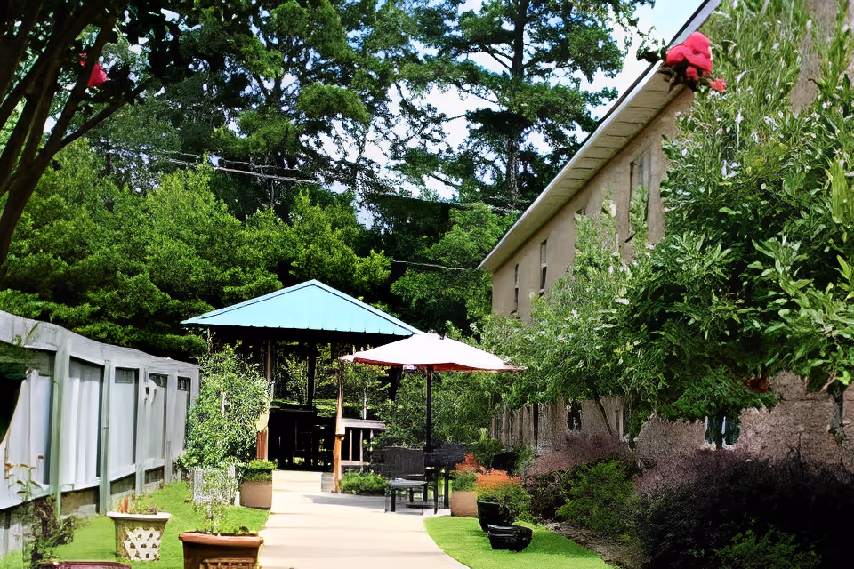 Outdoor pathway at Brandon Court Nursing facility with a gazebo featuring a blue roof, a patio umbrella, chairs, and lush green trees and bushes surrounding the area.